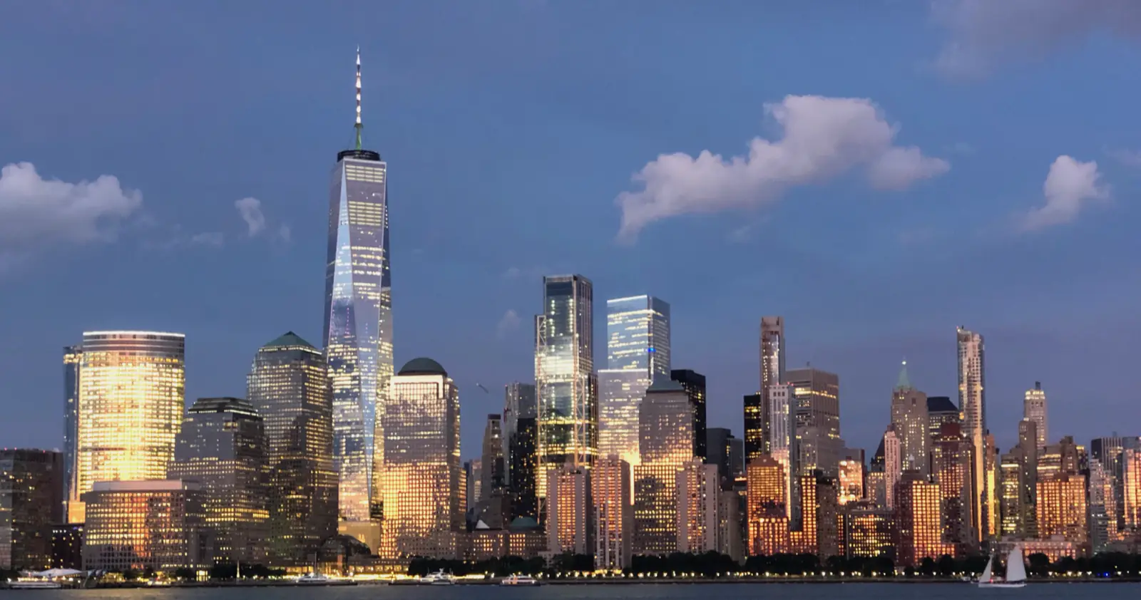 The Lower Manhattan skyline at dusk, lit gold by the setting sun, seen from across the Hudson River.